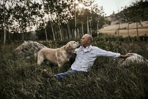 Bald man sits in a grassy field with a Labrador Retriever, which is licking his face. Trees and a hillside are in the background.