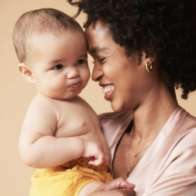 Portrait of a happy dark-skinned adult female holding an adorable infant with neutral expression in front of a plain tan background.