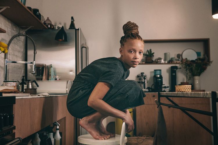 Young woman with braided hair in a topknot poses barefoot on a chair in a casual kitchen setting wearing a textured dark teal outfit.
