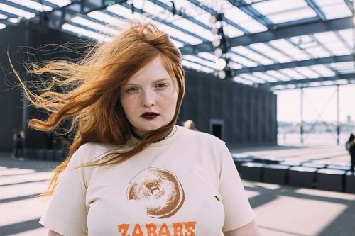 Portrait of a woman with red hair and a t-shirt with a donut graphic, posing in a large open space with a light gray floor.