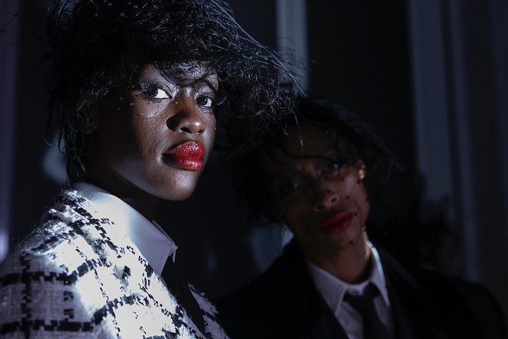 Two African-American women in dark suits with hats. The first woman has bright red lipstick and jewelry, while the second is blurry.