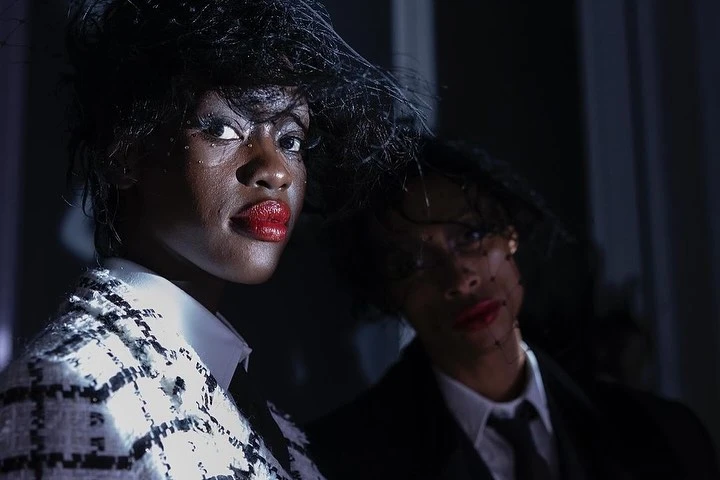 Two African-American women in dark suits with hats. The first woman has bright red lipstick and jewelry, while the second is blurry.