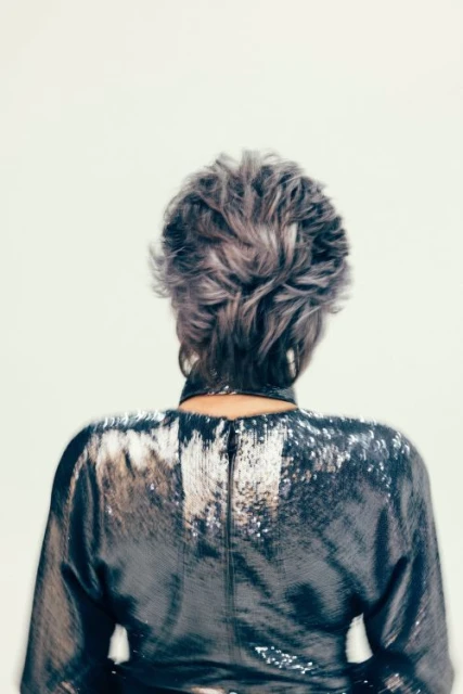 Back view of an adult female with short grey hair, wearing a shimmering long-sleeved dress against a plain white background, fashion shot.