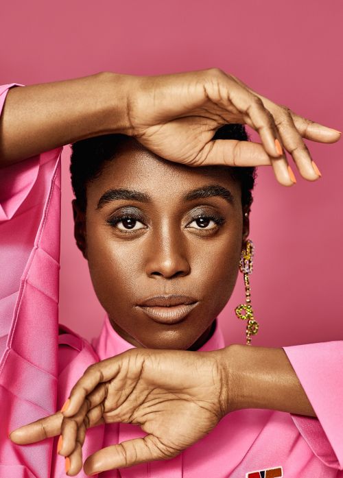 Close-up studio shot of an adult female with dark hair, wearing a pink shirt and long dangling earring against a pink background.