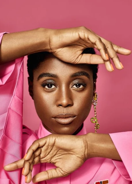 Close-up studio shot of an adult female with dark hair, wearing a pink shirt and long dangling earring against a pink background.