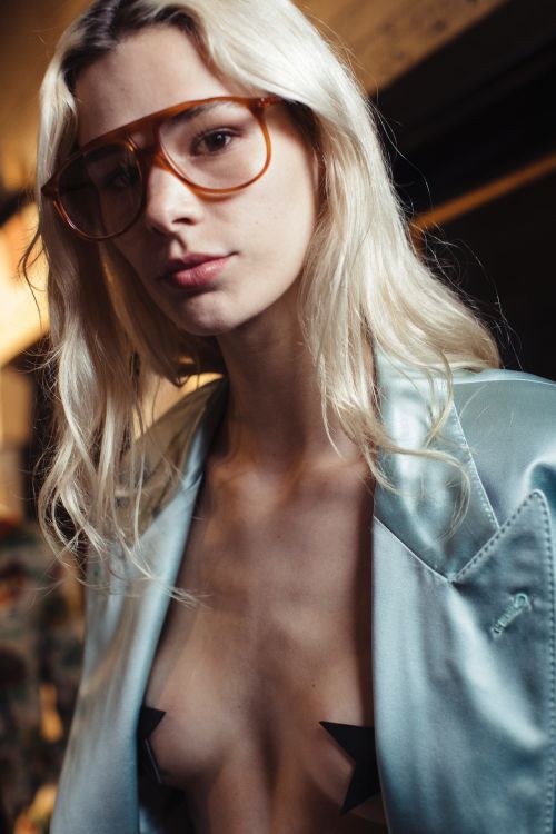 Portrait of a young adult female with blonde hair and aviator glasses, wearing a blazer with star-shaped nipple covers.