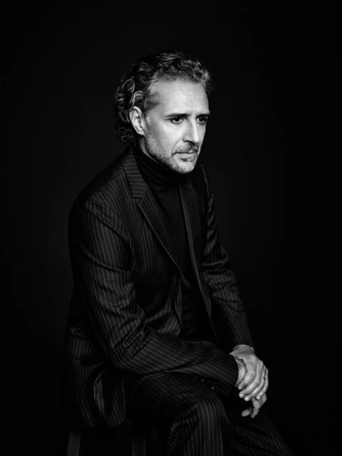 Black and white portrait of an adult male with curly hair, wearing a pinstripe suit and turtleneck, sitting on a stool against a dark background.