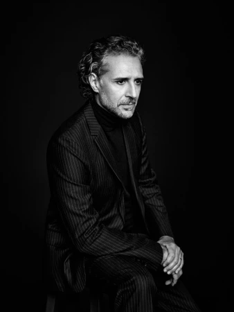 Black and white portrait of an adult male with curly hair, wearing a pinstripe suit and turtleneck, sitting on a stool against a dark background.