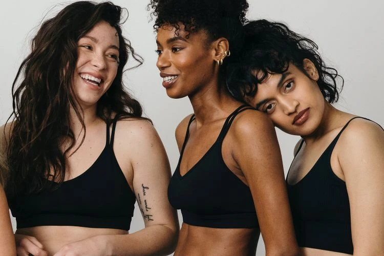 Three diverse women in black tops pose closely together against a light grey background, smiling slightly for a beauty or fashion shot.