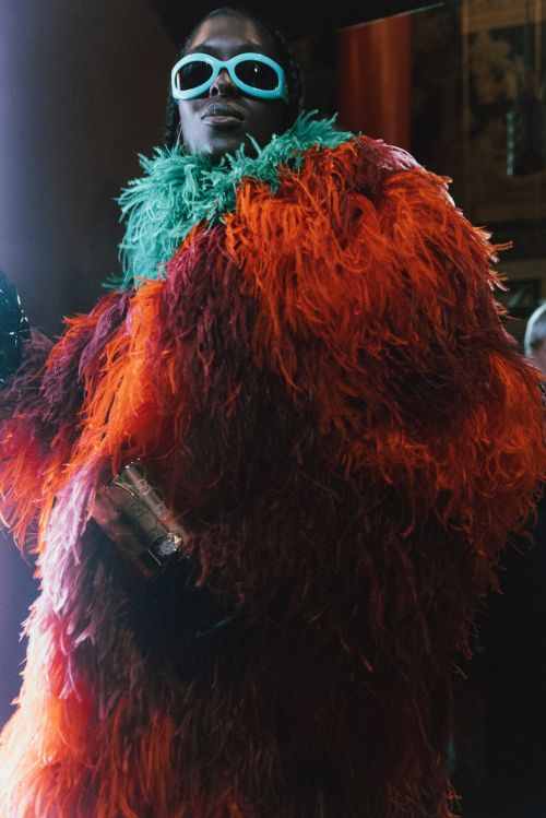 Adult wearing an orange feathered coat and light blue sunglasses poses indoors with blurred background in a portrait-style fashion shot.