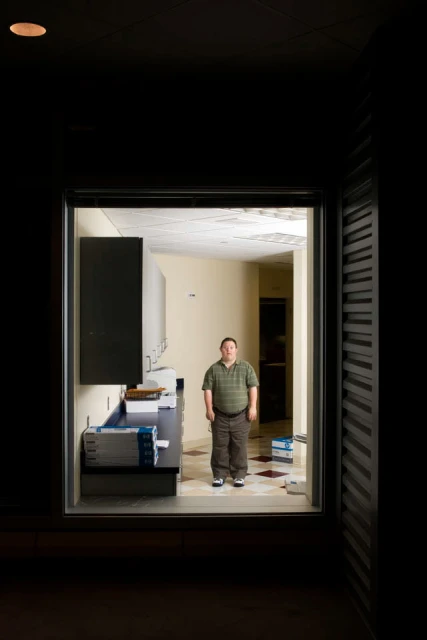 A man stands in an interior doorway of an abandoned-looking building, featuring a kitchen area with cabinets and a cluttered floor.