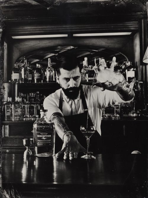 Monochromatic image of a bartender in a black apron pouring a drink from a mixing glass into a cocktail glass at a bar.