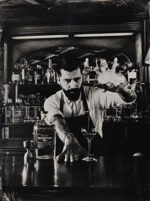 Monochromatic image of a bartender in a black apron pouring a drink from a mixing glass into a cocktail glass at a bar.