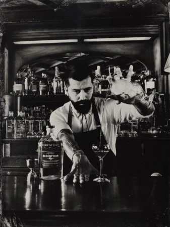 Monochromatic image of a bartender in a black apron pouring a drink from a mixing glass into a cocktail glass at a bar.