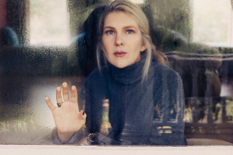 Woman in a dark turtleneck looking through a rain-streaked window with her hand pressed against the glass