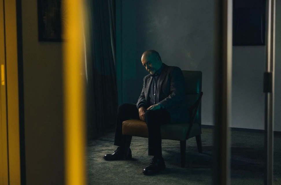 Mature man in a suit sits in a chair indoors, contemplative mood, teal and brown color palette, interior setting with framed pictures.