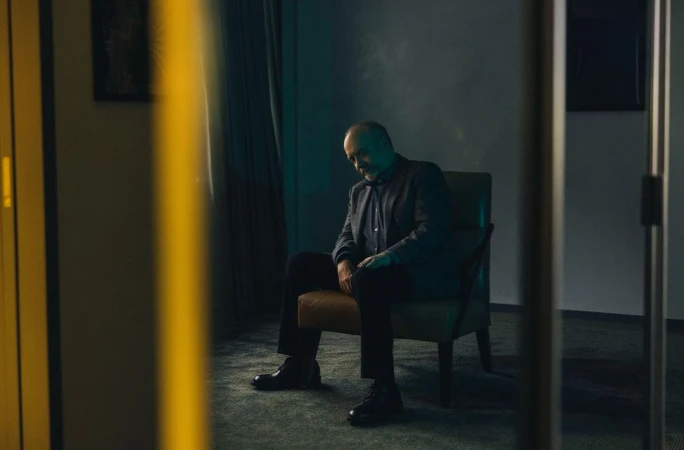 Mature man in a suit sits in a chair indoors, contemplative mood, teal and brown color palette, interior setting with framed pictures.