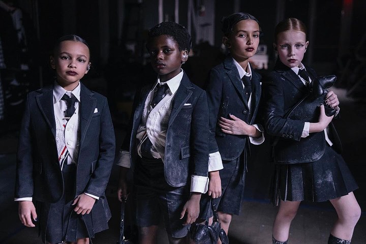 Four young children in dark blazers and skirts stand in a dim indoor setting with what appears to be light dust scattered on their attire.
