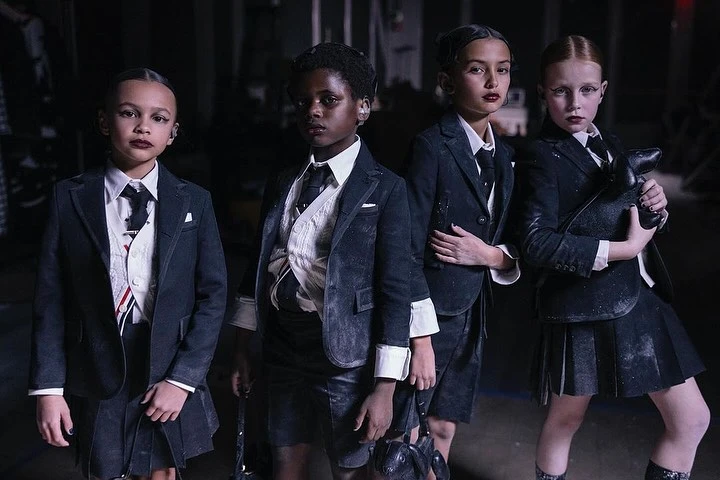 Four young children in dark blazers and skirts stand in a dim indoor setting with what appears to be light dust scattered on their attire.