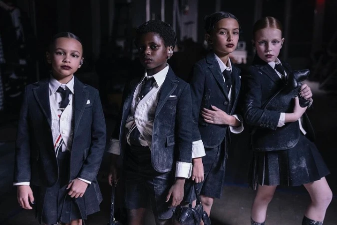 Four young children in dark blazers and skirts stand in a dim indoor setting with what appears to be light dust scattered on their attire.