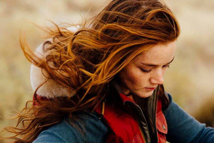 Close-up portrait of a woman with long red hair wearing a red vest outdoors, looking down with a neutral expression in a windy setting.