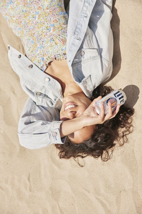 A woman with curly hair smiles on a sandy beach, holding a can of Miller Lite beer. She's wearing a denim jacket and floral dress.