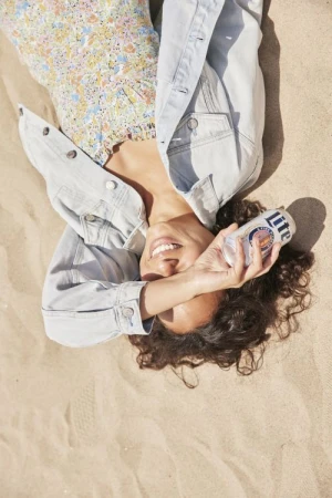 A woman with curly hair smiles on a sandy beach, holding a can of Miller Lite beer. She's wearing a denim jacket and floral dress.