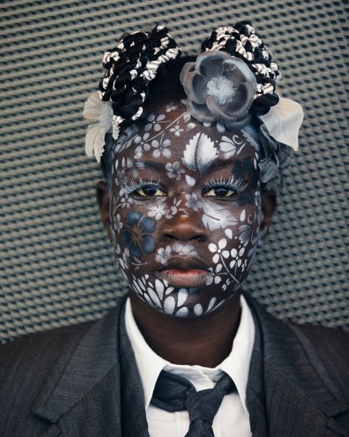 Portrait of Black woman with floral face paint, wearing a grey suit and tie against a textured background, creative makeup look.