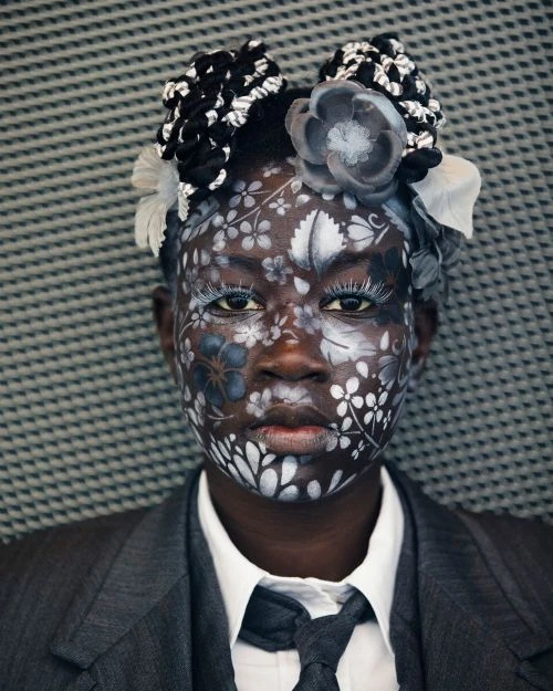 Portrait of Black woman with floral face paint, wearing a grey suit and tie against a textured background, creative makeup look.