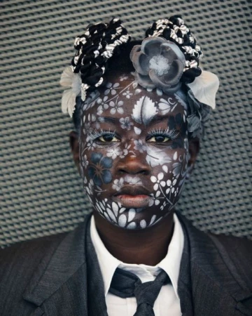 Portrait of Black woman with floral face paint, wearing a grey suit and tie against a textured background, creative makeup look.