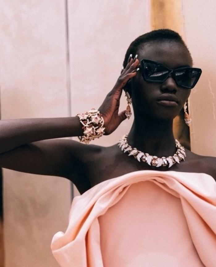 Elegant dark-skinned woman wearing oversized sunglasses, pink strapless dress, necklace and jewelry posing in an indoor setting.