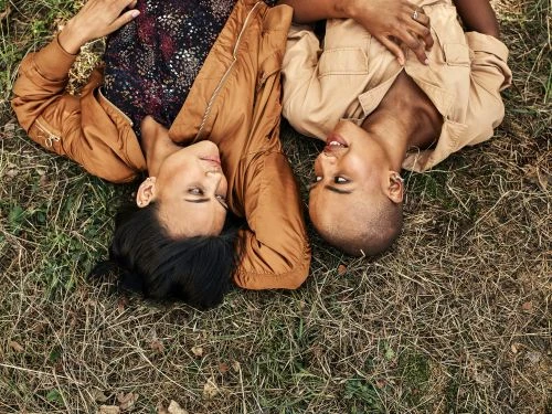 Two women lying head-to-head in dry grass, one with short hair wearing a tan shirt, the other with dark hair wearing a floral top and brown jacket.