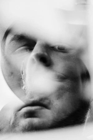 Monochrome portrait of a fair-skinned adult male's face with a small beard, blurred motion, and stark lighting creating multiple overlaid images.