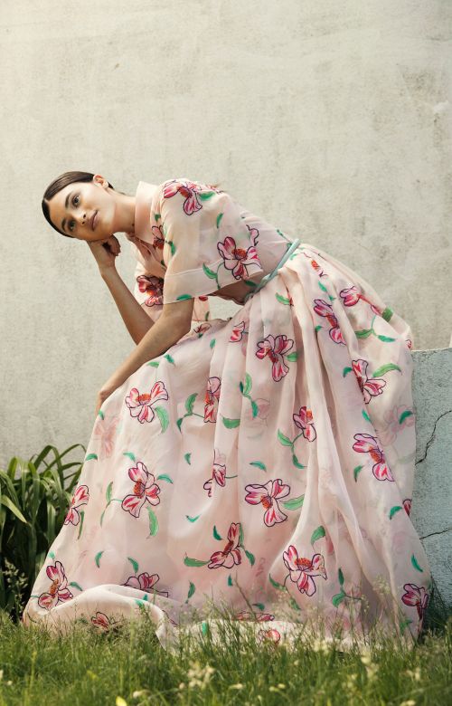 Adult woman in a long, light pink floral dress sitting outdoors in lush green grass next to a weathered concrete wall in daylight.