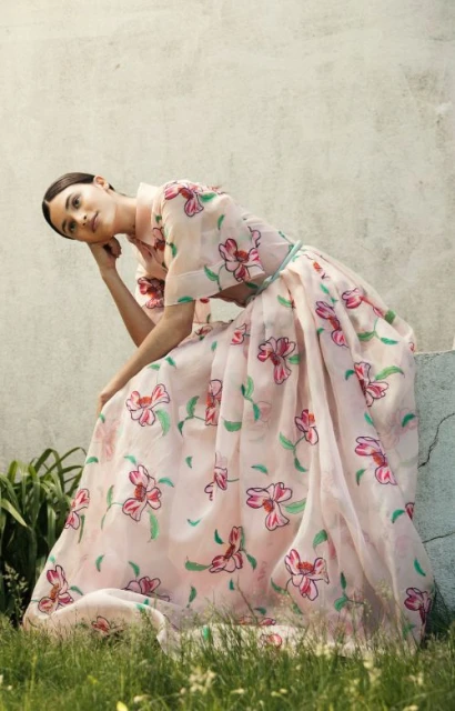 Adult woman in a long, light pink floral dress sitting outdoors in lush green grass next to a weathered concrete wall in daylight.