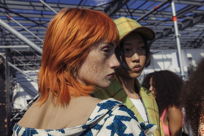 Two young women at an outdoor fashion event. One has bright red hair with freckles, the other is wearing a yellow plaid hat.