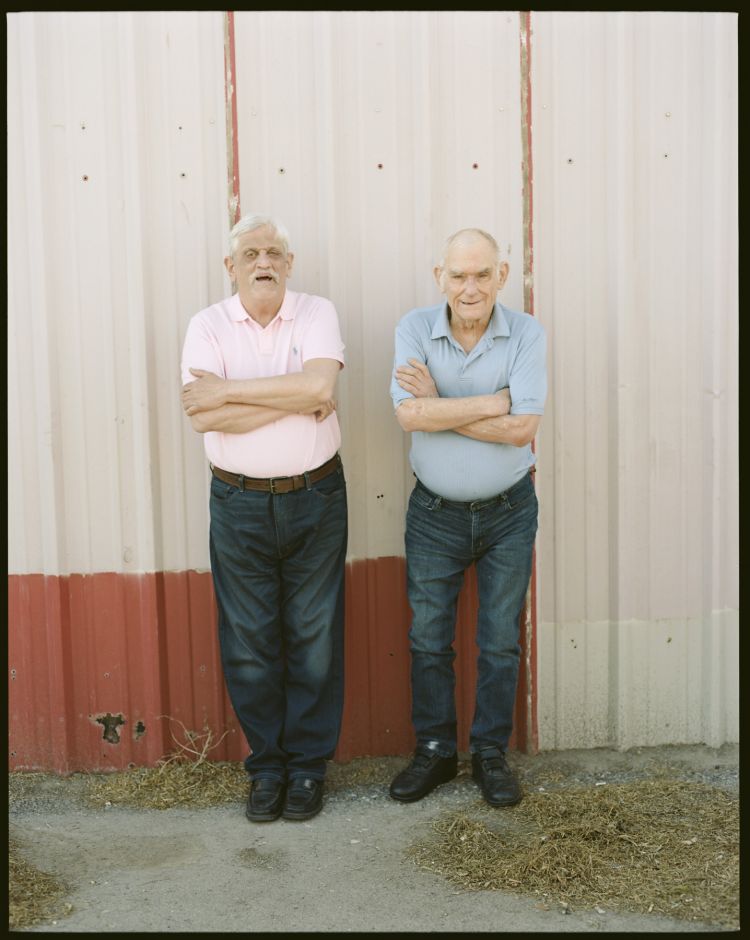 Two elderly men stand outdoors with arms crossed in front of a corrugated metal wall, wearing collared shirts and dark jeans in bright sunlight.