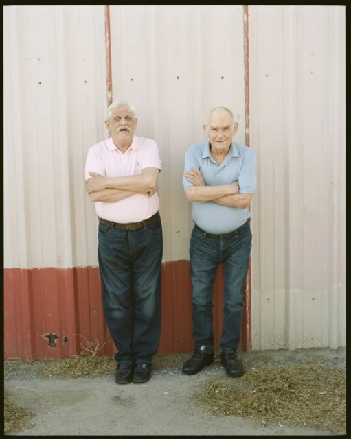 Two elderly men stand outdoors with arms crossed in front of a corrugated metal wall, wearing collared shirts and dark jeans in bright sunlight.