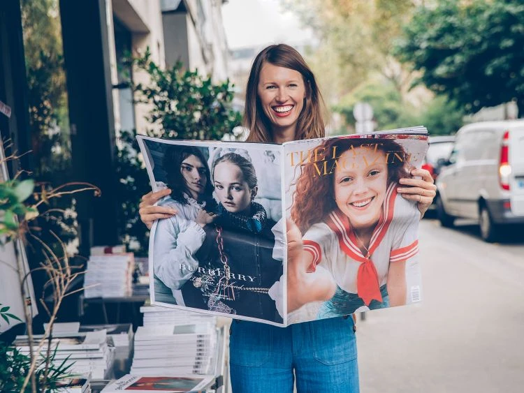 Woman smiles holding open fashion magazine with Burberry and 'The Last Human in the World' covers on city sidewalk.