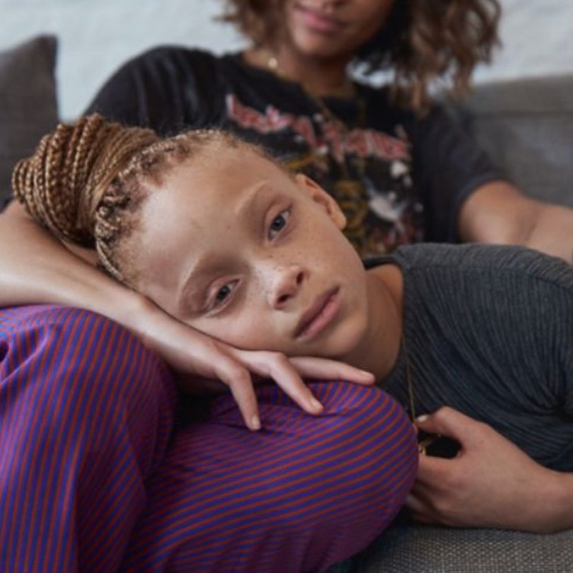 Child with braided hair rests head on striped pants of seated adult in gray shirt with graphic t-shirt in an indoor setting.