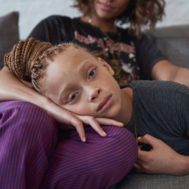 Child with braided hair rests head on striped pants of seated adult in gray shirt with graphic t-shirt in an indoor setting.
