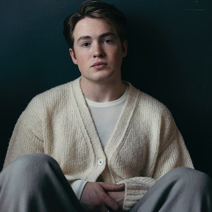 Portrait of actor Kit Connor wearing a cream knit cardigan against a dark background, photographed by Victoria Will.