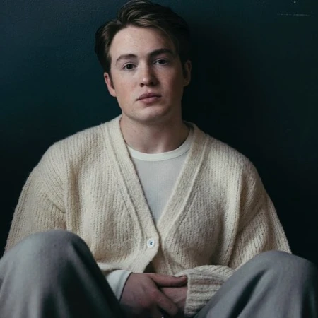 Portrait of actor Kit Connor wearing a cream knit cardigan against a dark background, photographed by Victoria Will.