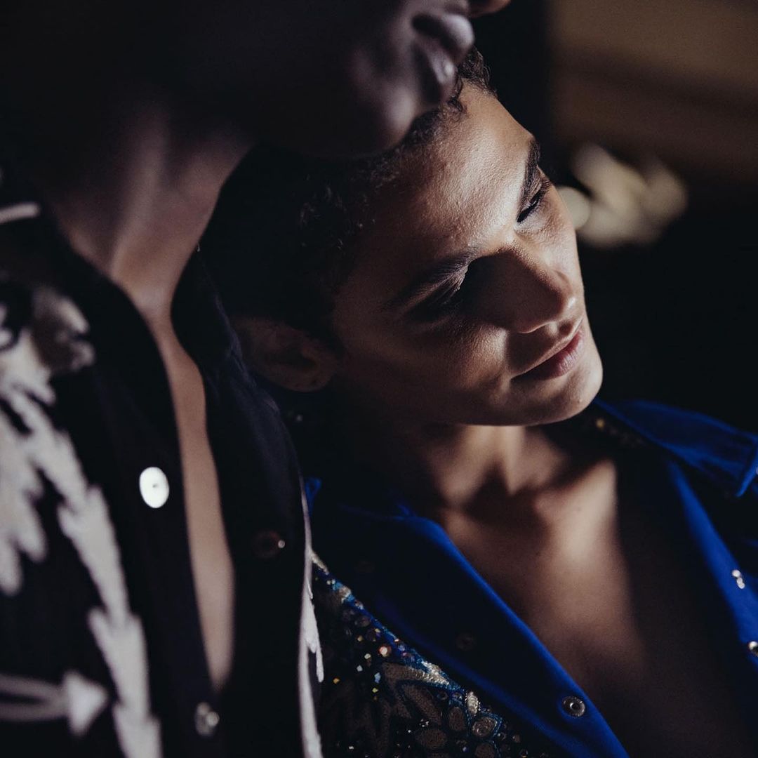 Close-up of two adults in dark indoor setting. One human is facing forward in blue embellished jacket, the other is leaning into frame.