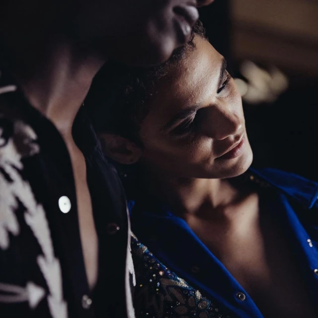 Close-up of two adults in dark indoor setting. One human is facing forward in blue embellished jacket, the other is leaning into frame.