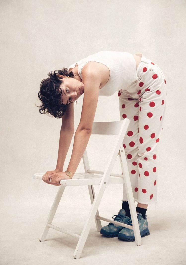 Woman with curly dark hair bends at the waist with her hands on a white chair. She wears a white top and red polka dot pants.
