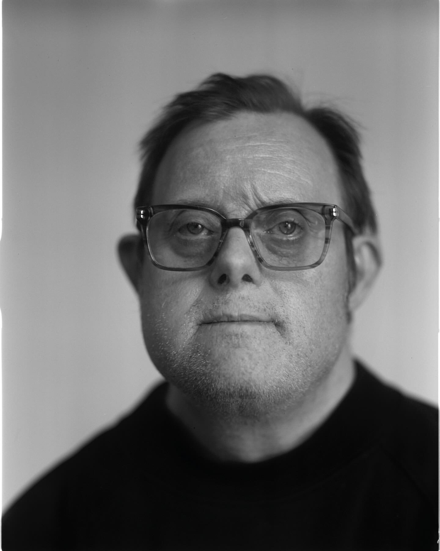 Monochrome portrait of a man with glasses in a black sweater against a light background, face forward, head and shoulders shot.