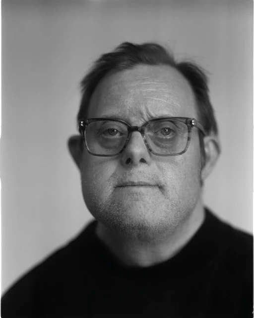 Monochrome portrait of a man with glasses in a black sweater against a light background, face forward, head and shoulders shot.