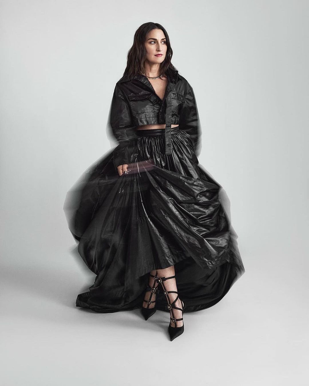 Woman in black crop top and voluminous black skirt posing against a white studio background with motion blur effect.