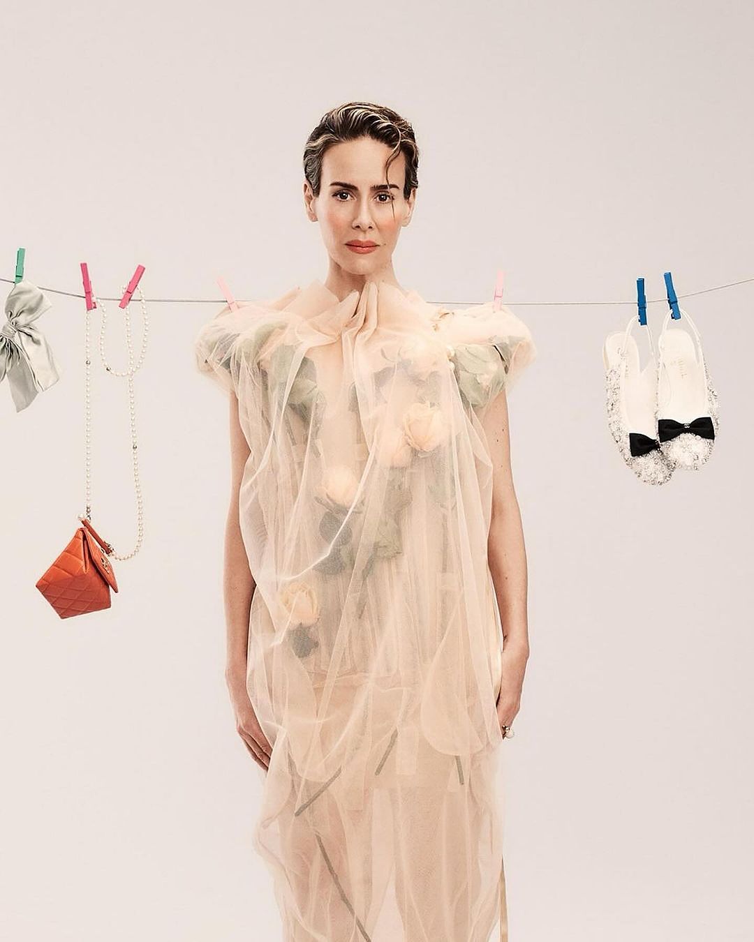 Woman in sheer gown poses in a photo studio, surrounded by fashion accessories hanging from clothespins and string against a light backdrop.